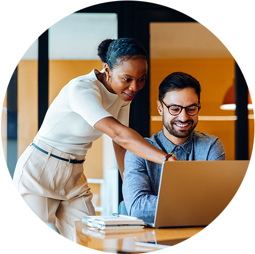 Picture of a person pointing at a laptop screen as someone is sitting on the chair in front of the laptop smiling wearing glasses.