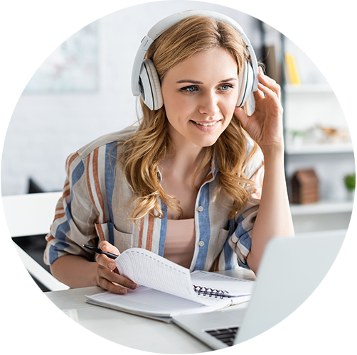 A person smiling looking at their laptop screen, wearing over the head headphones, also making notes in a notepad